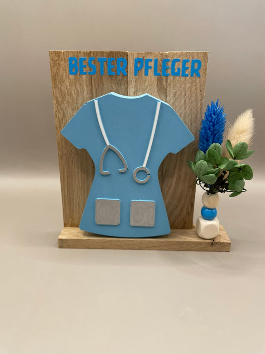 Geschenk in Form eines Kittels in blau mit der Überschrift "Bester Pfleger". Dekoriert mit Trockenblumen, alles auf einem Holzaufsteller.