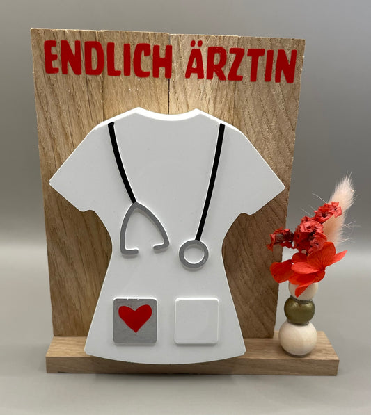 Geschenk in Form eines Kittels in weiß mit der Überschrift "Endlich Ärztin". Dekoriert mit Trockenblumen, alles auf einem Holzaufsteller.